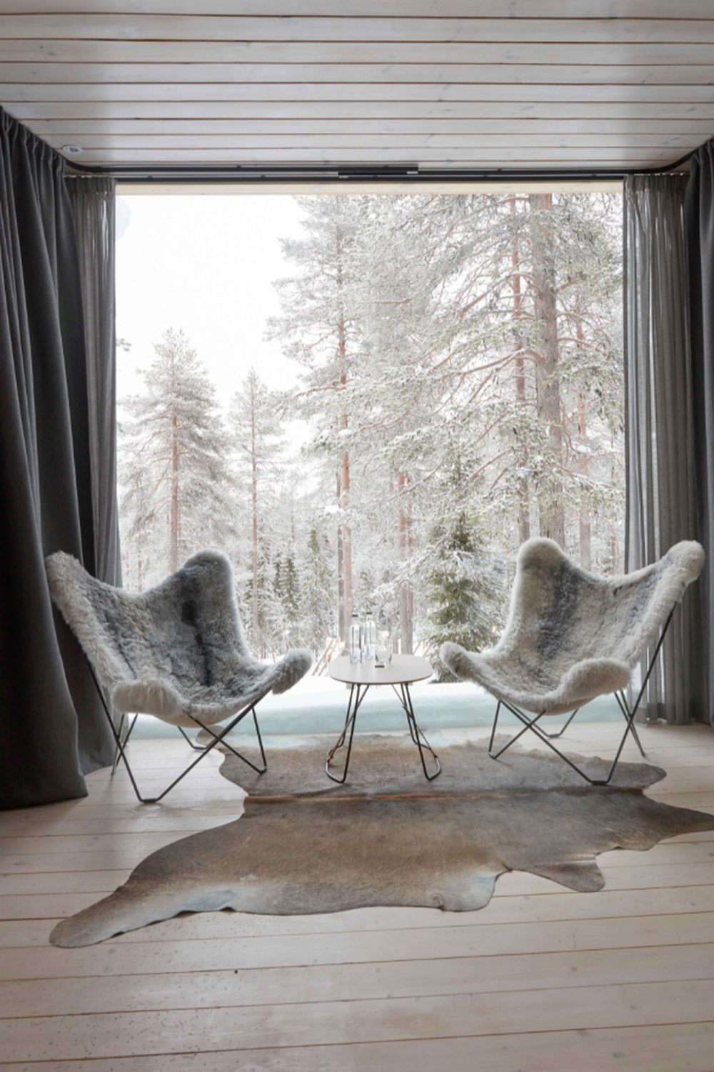 coffee table between two fur butterfly chairs in ski chalet with animal skin rug, snowy forest with pines in background